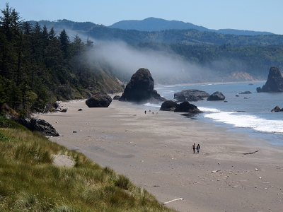 [A wide sand expanse with a few huge black rocks and some people walking on the sand as the waves roll in. At the near edge is a grassy expanse while the far edge is treed. An expanse of fog partially obscures the view of the far trees.]